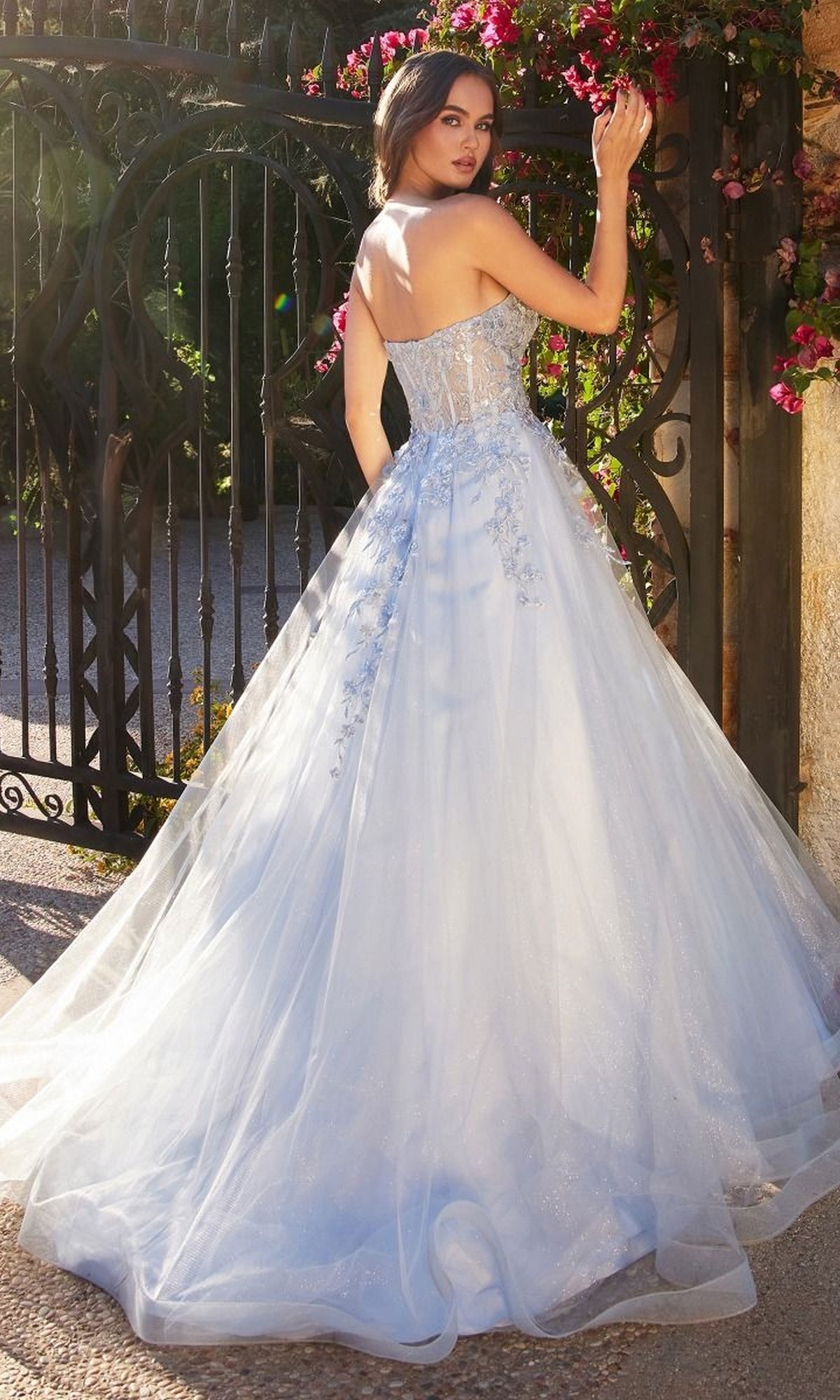 Wearing the Pastel Blue Ball Gown A1339 by Andrea and Leo, a woman stands outdoors near a black iron gate, turning to look over her shoulder.